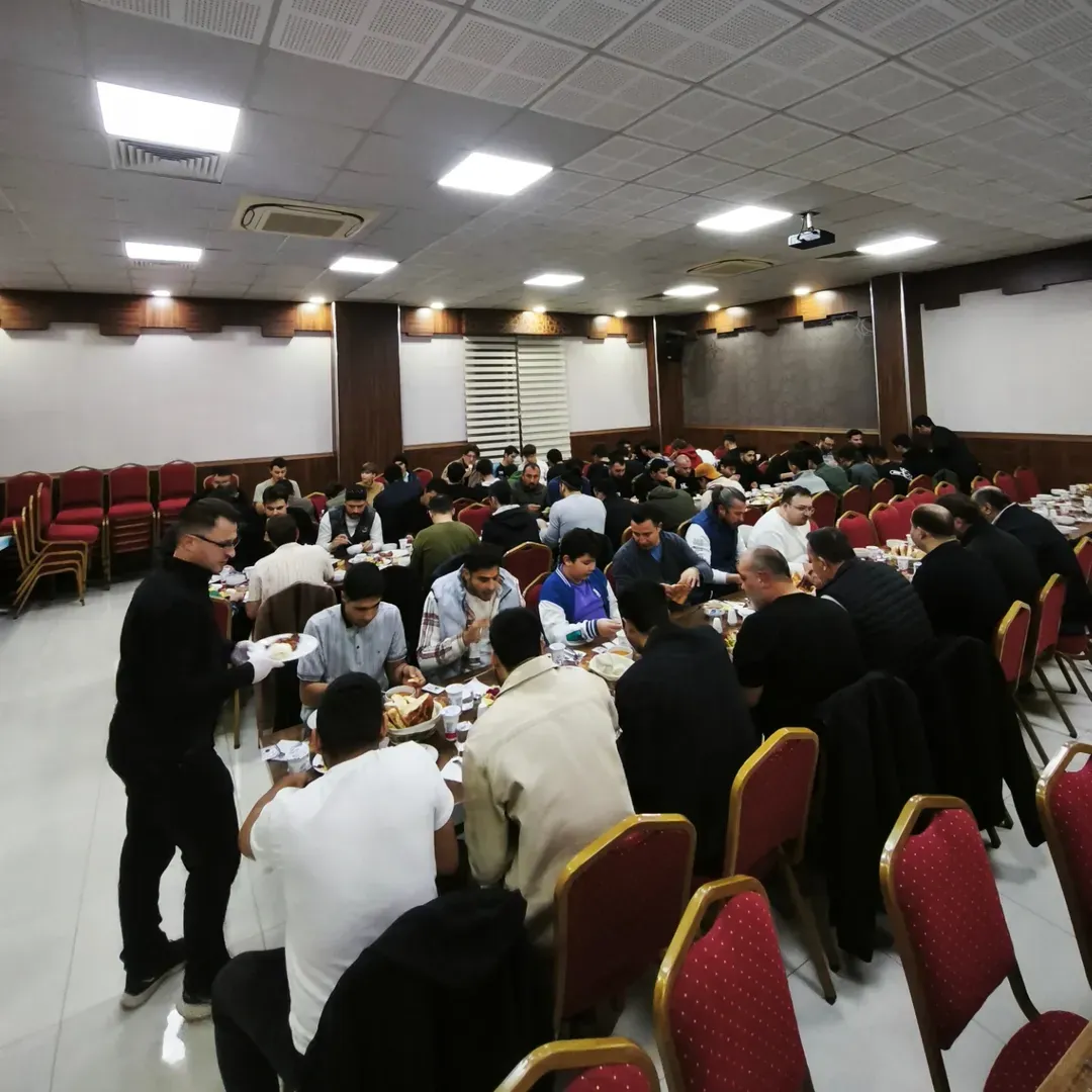 Geniş bir salonda uzun sofralarda toplu yemek yiyen kalabalık topluluk